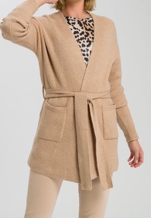 Femme portant un cardigan en tricot beige noué à la taille sur un haut à imprimé léopard et un pantalon beige, avec une main levée près de sa tête.