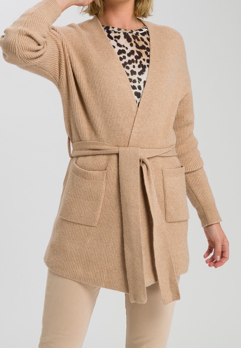 Kvinde iført en beige strikket cardigan, der er bundet om taljen over en leopardmønstret top og beige bukser, med den ene hånd hævet nær sit hoved.