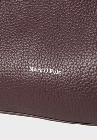 Texture en cuir bordeaux avec une finition granuleuse, présentant le logo blanc embossé "Marc O'Polo" centré sur la surface.