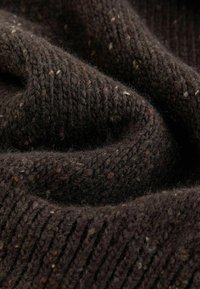 Dunkelbrauner Strickstoff mit gerippter Textur und kleinen Farbflecken in helleren Tönen. Das Material wirkt weich und leicht fusselig.