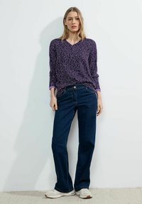 Blusa stampata viola con scollo a V e maniche lunghe, abbinata a jeans dritti blu scuro e sneakers bianche.
