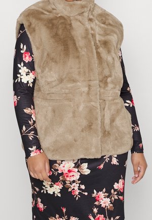 Beige faux fur ærmeløs vest båret over en sort kjole med pink og hvide blomsterprint.