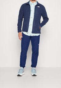 Man i marinblå fleecetröja över ljusblå skjorta med djurtryck, marinblå byxor och ljusblå sneakers står mot vit bakgrund.