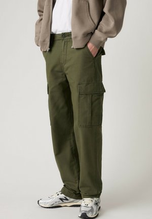 Persona vestida con pantalones cargo verde oliva, chaqueta beige con cremallera, camisa blanca y zapatillas New Balance blancas y grises, de pie con las manos en los bolsillos.