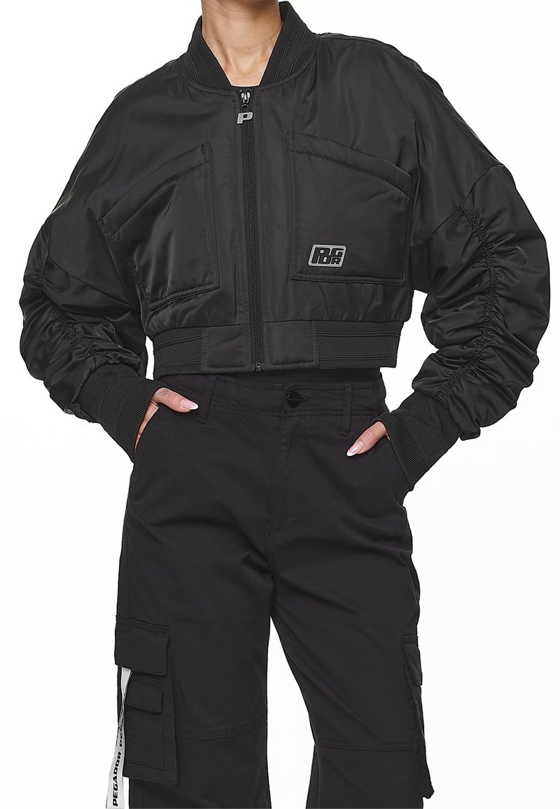 Femme portant une veste bomber noire courte à manches froncées et un pantalon cargo noir taille haute avec des détails de poches.