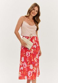 Květinový top bez rukávů v jemné růžové barvě spárovaný s červenou midi sukní s velkými růžovými a bílými květinovými vzory; drží béžovou kabelku.