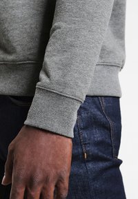 Felpa grigia con polsini a costine, realizzata in morbido tessuto di cotone. Presenta una finitura strutturata ed è abbinata a jeans in denim scuro.