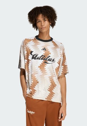 Persona con cabello rizado que lleva una camiseta Adidas marrón y blanca con patrón de zigzag y pantalones Adidas marrones, de pie con una mano en el bolsillo.