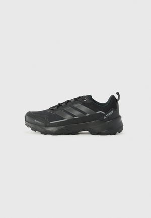 Chaussure de sport basse noire avec semelle texturée, système de laçage, et bandes et logos de marque sur le côté, conçue pour les activités en extérieur.
