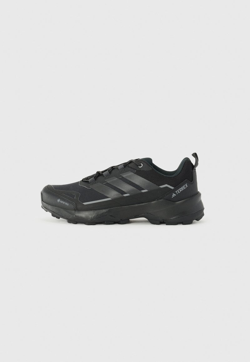 Chaussure de sport basse noire avec semelle texturée, système de laçage, et bandes et logos de marque sur le côté, conçue pour les activités en extérieur.