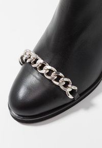 Botte en cuir noir à bout pointu, ornée d'une chaîne argentée sur le devant. Texture lisse avec une semelle plate.