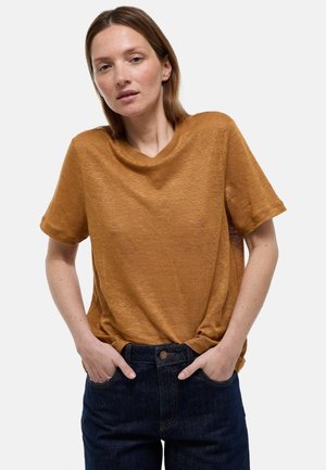 Femme à la peau claire et aux cheveux bruns raides porte une chemise marron ample à manches courtes et un jean bleu foncé, mains dans les poches, fond neutre.