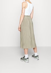 Person der står med ryggen til, iført en hvid ærmeløs top, en beige plisseret midi-nederdel, hvide sokker og grønne-sorte sportssko.