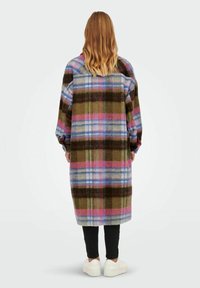 Woman with wavy blonde hair wearing a long multicolor plaid coat, black pants, and white sneakers, standing facing away on a plain background.