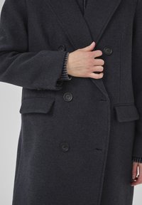 Asmuo užsisega mygtukus ant tamsiai pilkos vilnonės dvigubos eilės paltos su kišenėmis ir matomomis raštuotomis megztinėmis rankovėmis.