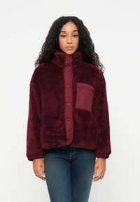Bordeaux fleece-takk med høy krage, trykknapper og en lomme på venstre bryst. Har en myk, teksturert overflate og avslappet passform.