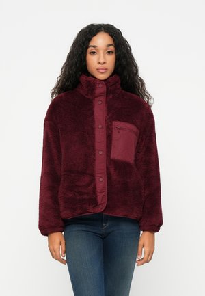 Bordeaux fleece jack met hoge kraag, drukknoopsluiting en een borstzak aan de linkerkant. Heeft een zachte, gestructureerde oppervlakte en een comfortabele pasvorm.