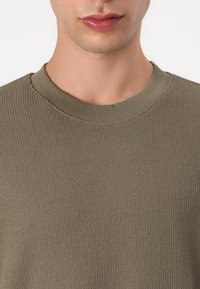 Pulover tricotat în verde măsliniu, cu guler rotund. Textura materialului este moale și ușor elastică, având cusături detaliate în jurul gâtului.