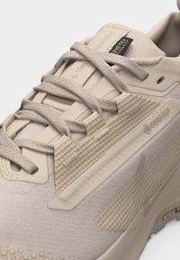 Artimas smėlio spalvos Gore-Tex sportbačio su tekstūruotais intarpais ir tvirtai surištomis raištelėmis vaizdas ant balto fono.