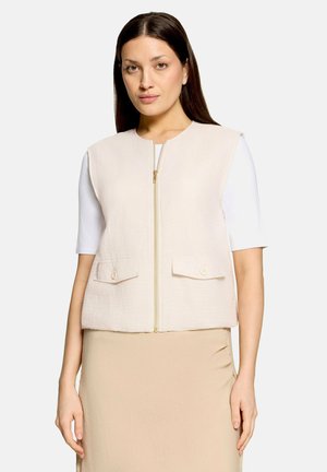 Femme aux cheveux lisses et foncés portant un gilet beige sans manches zippé par-dessus un haut blanc à manches courtes et une jupe beige sur un fond clair.