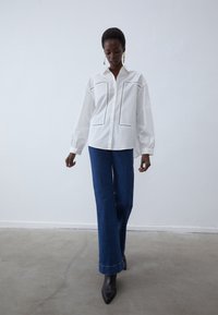 Witte langekouthemd met een gestructureerde kraag, twee borstzakken en contrasterende stiksels, gecombineerd met brede blauwe denim broek.