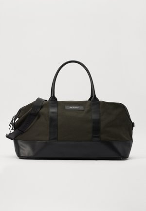 Sac de sport en toile verte avec des accents en cuir noir, forme arrondie, poignées doubles et bandoulière réglable avec un texte embossé.