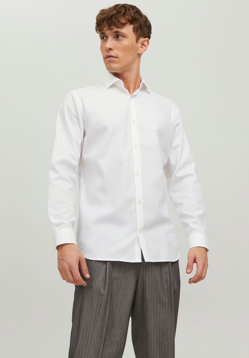 Giovane uomo che indossa una camicia bianca attillata abbottonata e pantaloni grigi a sottili righe, in piedi contro uno sfondo chiaro uniforme, che guarda verso sinistra.