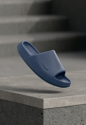 Pojedyncza niebieska klapka Nike unosząca się nad betonową powierzchnią, w tle rozmyte schody.