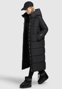 Langer, schwarzer Daunenmantel mit hohem Kragen und großer Kapuze. Verfügt über seitliche Taschen und einen Reißverschluss, kombiniert mit glänzenden, schwarzen Stiefeletten.