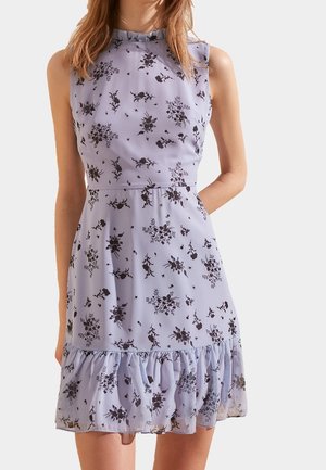 Robe lavande sans manches avec imprimé floral noir, col montant, taille ajustée et ourlet volanté portée par une personne debout.