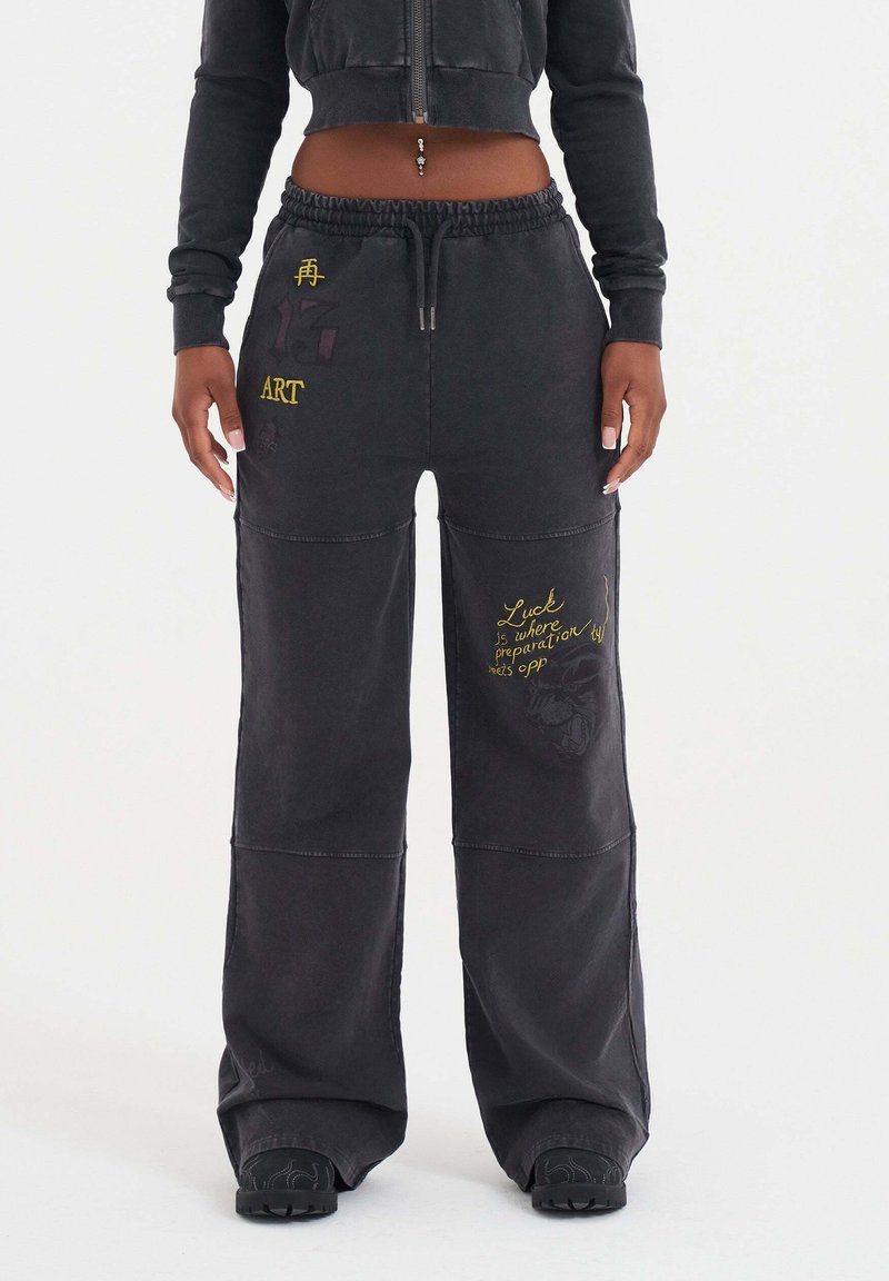 Svarta lösa sweatpants med dragsko i midjan, med gult och lila text och grafiska detaljer på benen och fickorna.