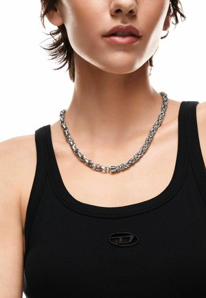 Close-up van een persoon die een zilverkleurige ketting draagt en een zwarte mouwloze top met een klein ovaal logo op de borst.