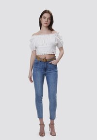 Top corto blanco sin mangas con detalles de encaje, combinado con jeans ajustados de color azul claro y un cinturón trenzado marrón claro. Sandalias con un tacón modesto.