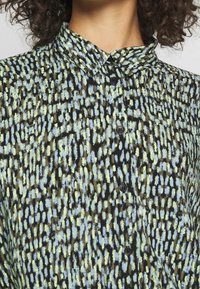 Detailní záběr na košili s límečkem s abstraktním vzorem štětcových tahů v černé, modré, zelené a béžové barvě a s černými knoflíky.