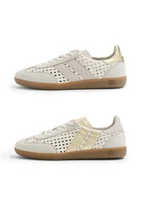 Deux baskets beiges avec des panneaux latéraux tissés, des lacets à l'avant, des semelles en gomme et des accents dorés brillants au talon et aux bandes, vues de l'extérieur.