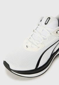 White athletic shoe featuring a textured mesh upper, black accents, and prominent logo detail on the tongue and outer side.