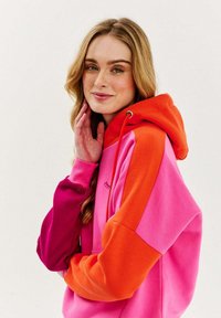 Barevná oversized mikina v růžové, fialové a oranžové barvě. Disponuje kapucí s tkaničkami, kontrastními rukávy a měkkou, texturovanou tkaninou.