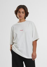 Tricou oversized alb, cu mâneci scurte și guler rotund. Prezintă text brodat roșu "Interzis" pe piept. Material moale din bumbac.