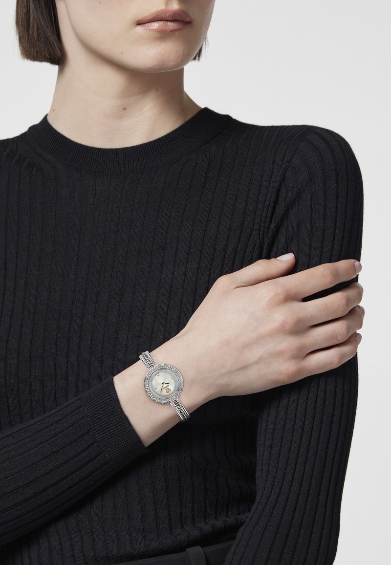 Kobieta z krótkimi włosami, ubrana w czarny sweter ze żłobieniami oraz srebrny zegarek na nadgarstku z ozdobną ramką i wzorzystym paskiem na lewej ręce.