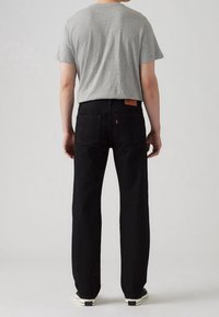 Homme portant un jean noir à jambe droite, un t-shirt gris à manches courtes et des baskets noir et blanc, debout de dos sur un fond uni.