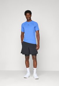 Homme debout portant un maillot Nike Pro bleu, un short noir superposé, des chaussettes blanches Nike et des baskets blanches sur un fond uni.