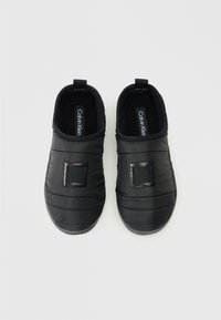 Zapatos negros sin cordones con una textura suave y acolchada. Presentan un parche de logo rectangular y un acabado liso, diseñados para la comodidad y facilidad de uso.