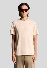 Licht perzik kleurig T-shirt met korte mouwen, gemaakt van zachte stof, met een subtiele geborduurde logo op de borst. Gekozen in combinatie met donkere zonnebril.