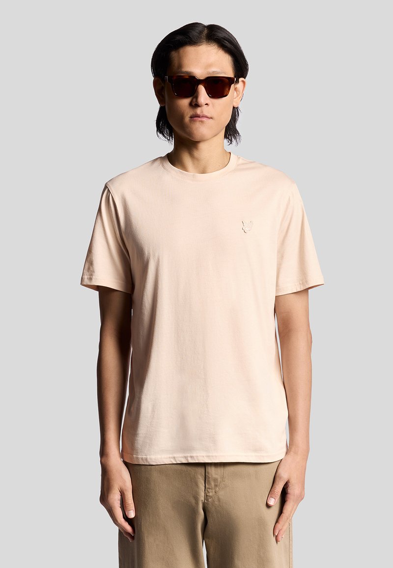 Licht perzik kleurig T-shirt met korte mouwen, gemaakt van zachte stof, met een subtiele geborduurde logo op de borst. Gekozen in combinatie met donkere zonnebril.