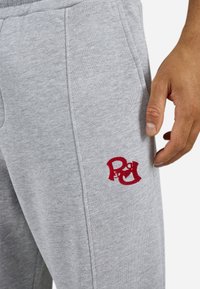 Nærbillede af grå sweatpants med rødt broderet logo nær lommen, ved siden af en persons højre hånd.