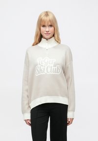 Világos bézs színű cipzáros pulóver fehér bordás mandzsettákkal és szegéllyel. Az elülső részen puha fehér betűkkel "Le Ger Ski Club" felirattal. Puha anyagból.