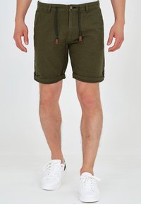Shorts en lin verts avec poches avant, cordon tressé et fermeture à bouton. Ourlets retroussés et détails de boutons sur les côtés. Portés avec des baskets blanches.