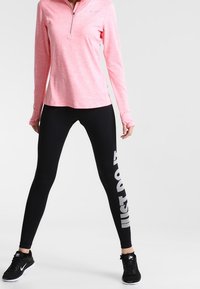 Różowy długi rękaw z zamkiem, w połączeniu z czarnymi legginsami z białym napisem "NIKE". Obie odzież wykonana jest z elastycznego materiału.