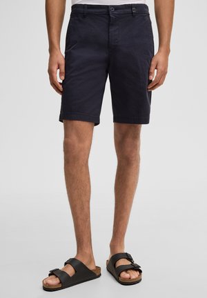 Mann trägt marineblaue kniehohe Shorts und schwarze Sandalen mit Schnalle, steht vor einem schlichten Hintergrund.