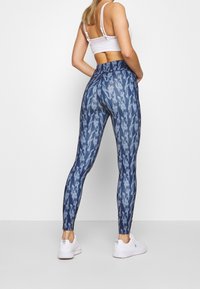 Modré legíny s vzorem vyrobené z elastické látky, s vysokým pasem a těsným střihem, v kombinaci s bílými sportovními botami.